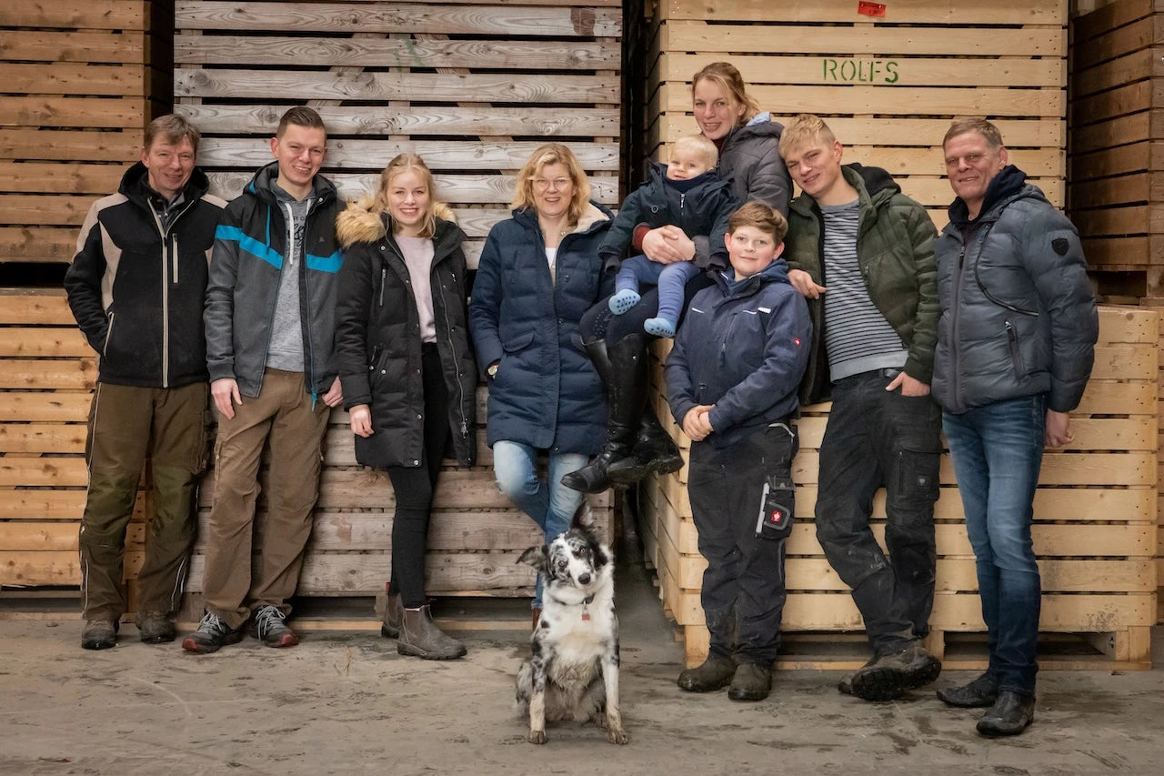 Eine Familie mit neun Personen und einem Hund posiert fröhlich in einer Halle vor gestapelten Holzkisten. Die Erwachsenen und Kinder tragen warme Winterkleidung. Eine Frau hält ein Kleinkind auf dem Arm. Der Hund sitzt im Vordergrund in der Mitte.